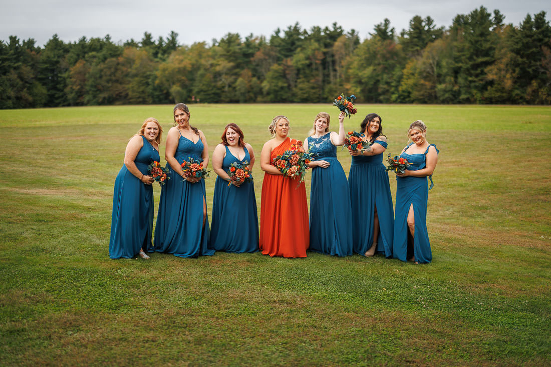 Bliss Farm Wedding-8 A group of people outdoors in a grassy field at Crestview Country Club, wearing formal dresses; one in orange and the others in blue, capturing stunning wedding photos amidst nature's beauty.