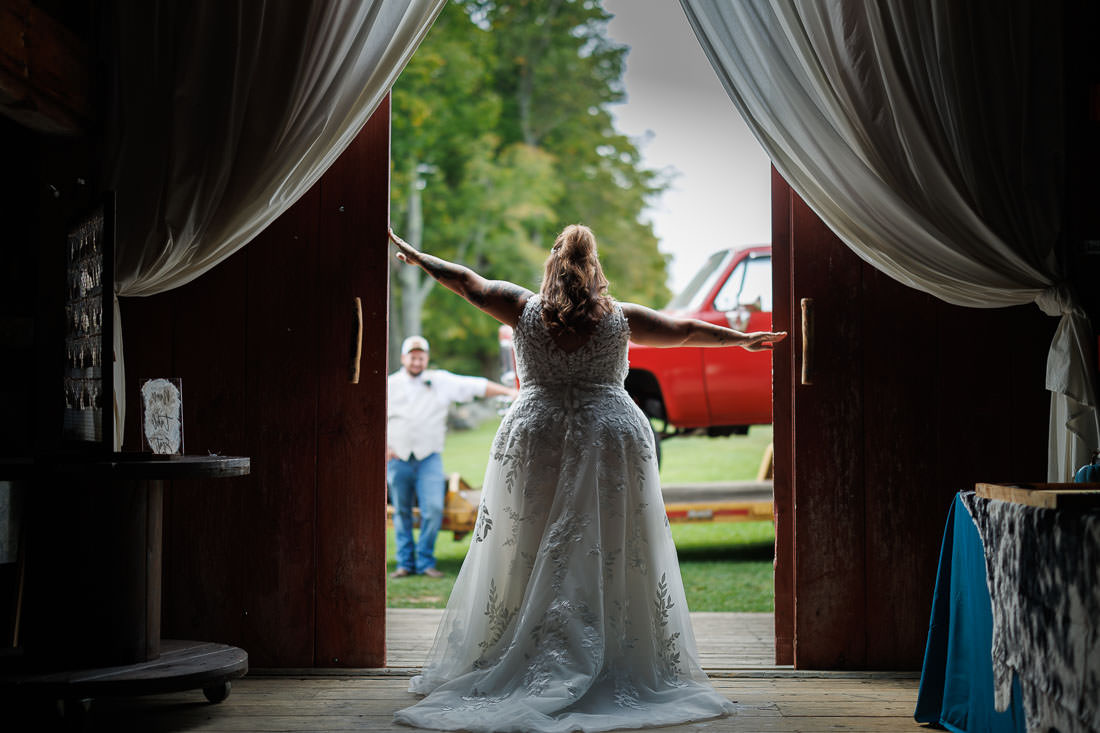 Bliss Farm Wedding-9 A bride in a white dress stands with arms outstretched in a doorway at Crestview Country Club, looking outside, where a man and a red truck are visible.