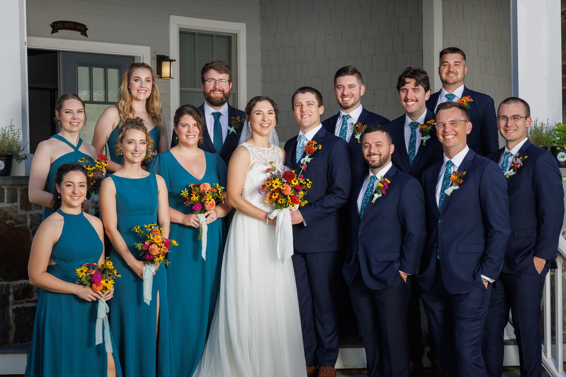 A bride and groom stand with a group of formally dressed individuals, including bridesmaids in teal dresses and groomsmen in suits, all holding flowers, posing on a porch at Bliss Farm.