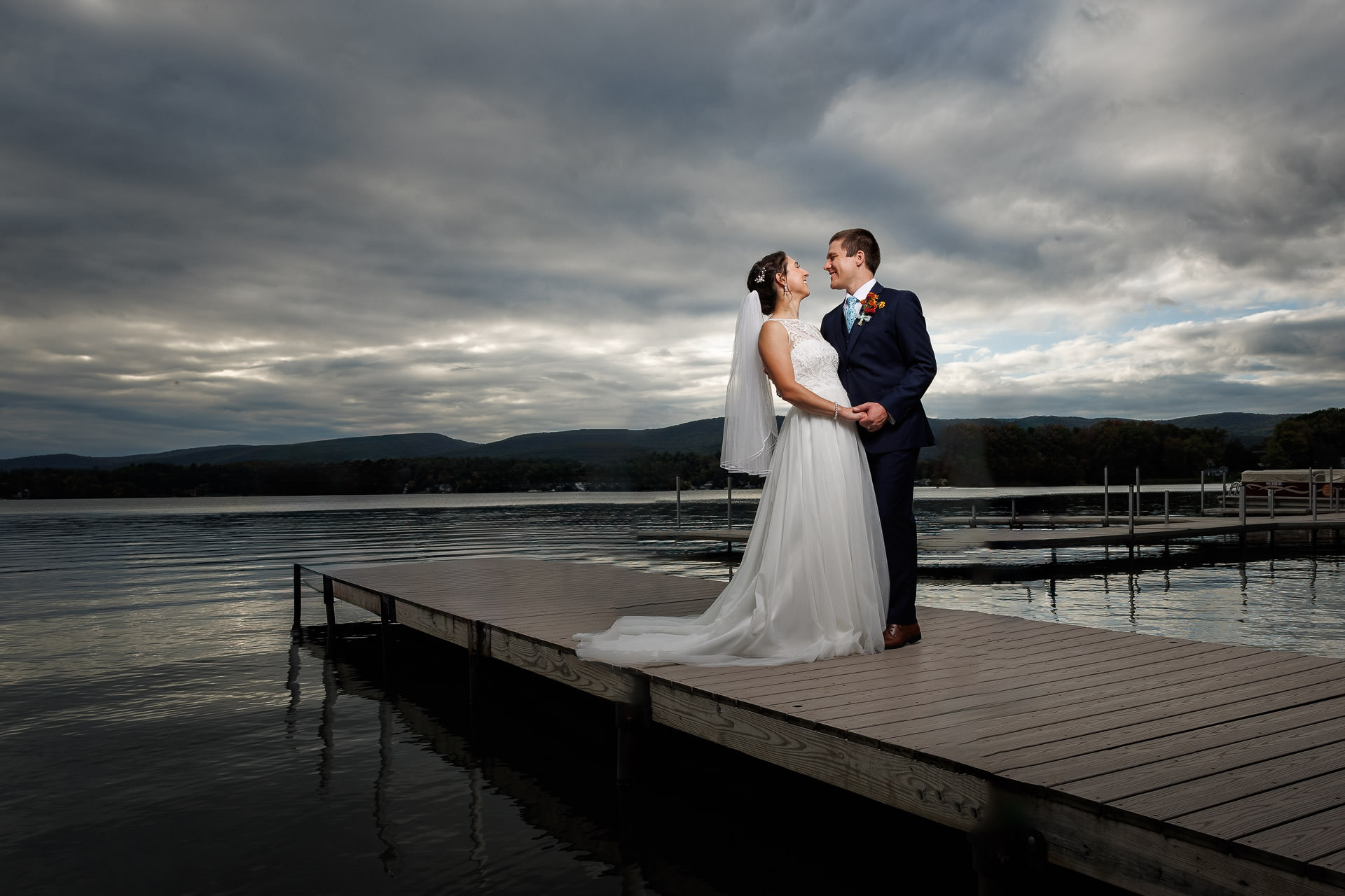 The bride and groom share a tender moment on a wooden dock by the lake, their hands intertwined as they gaze into each other's eyes beneath a cloudy sky—a serene capture from their Bliss Farm wedding photos.