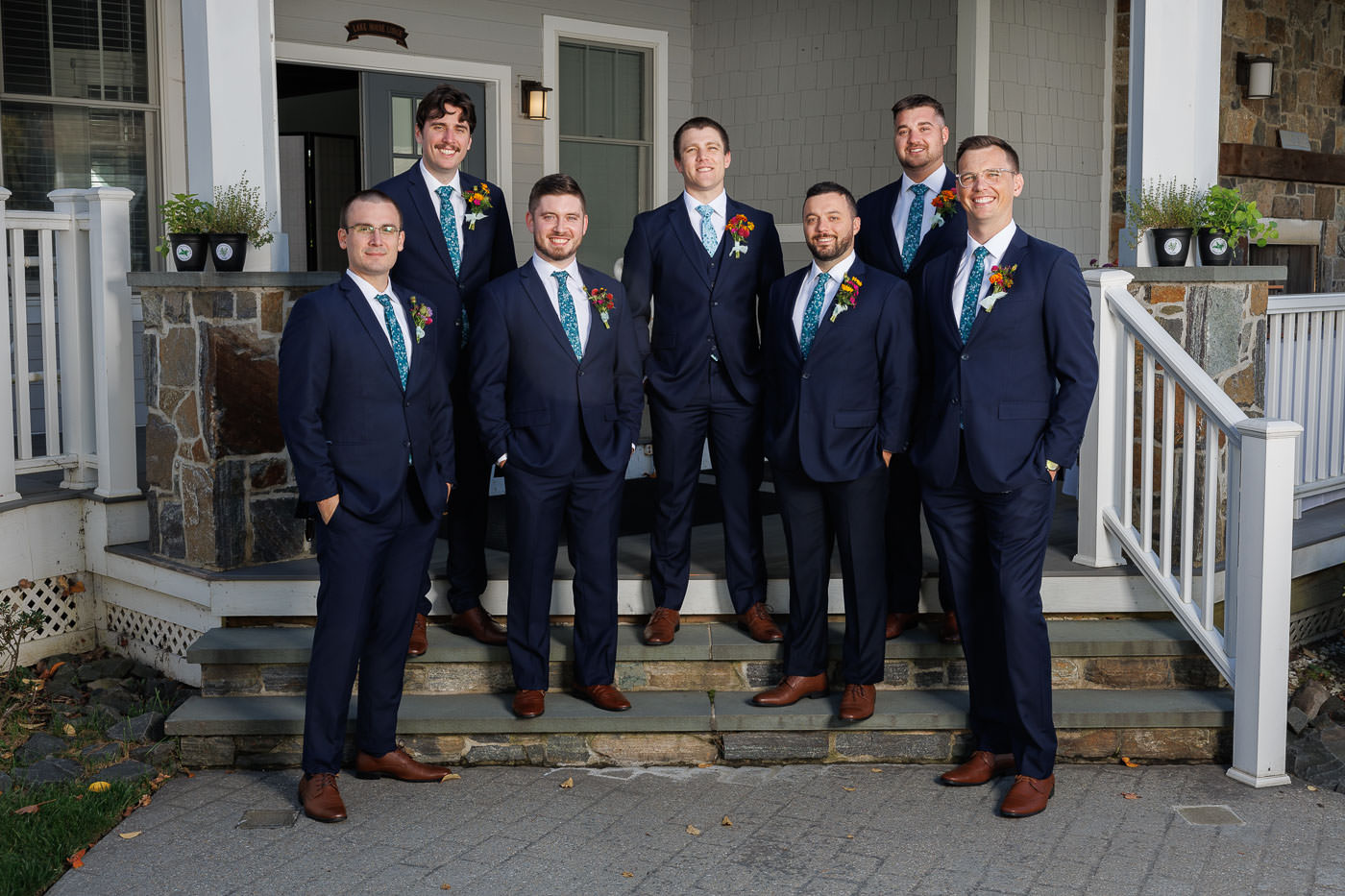 Lake house guest cottages wedding-10 Seven men in dark suits with floral boutonnieres stand on the porch steps of a charming farmhouse. The scene captures the elegance and camaraderie of a Bliss Farm wedding, creating picture-perfect memories.