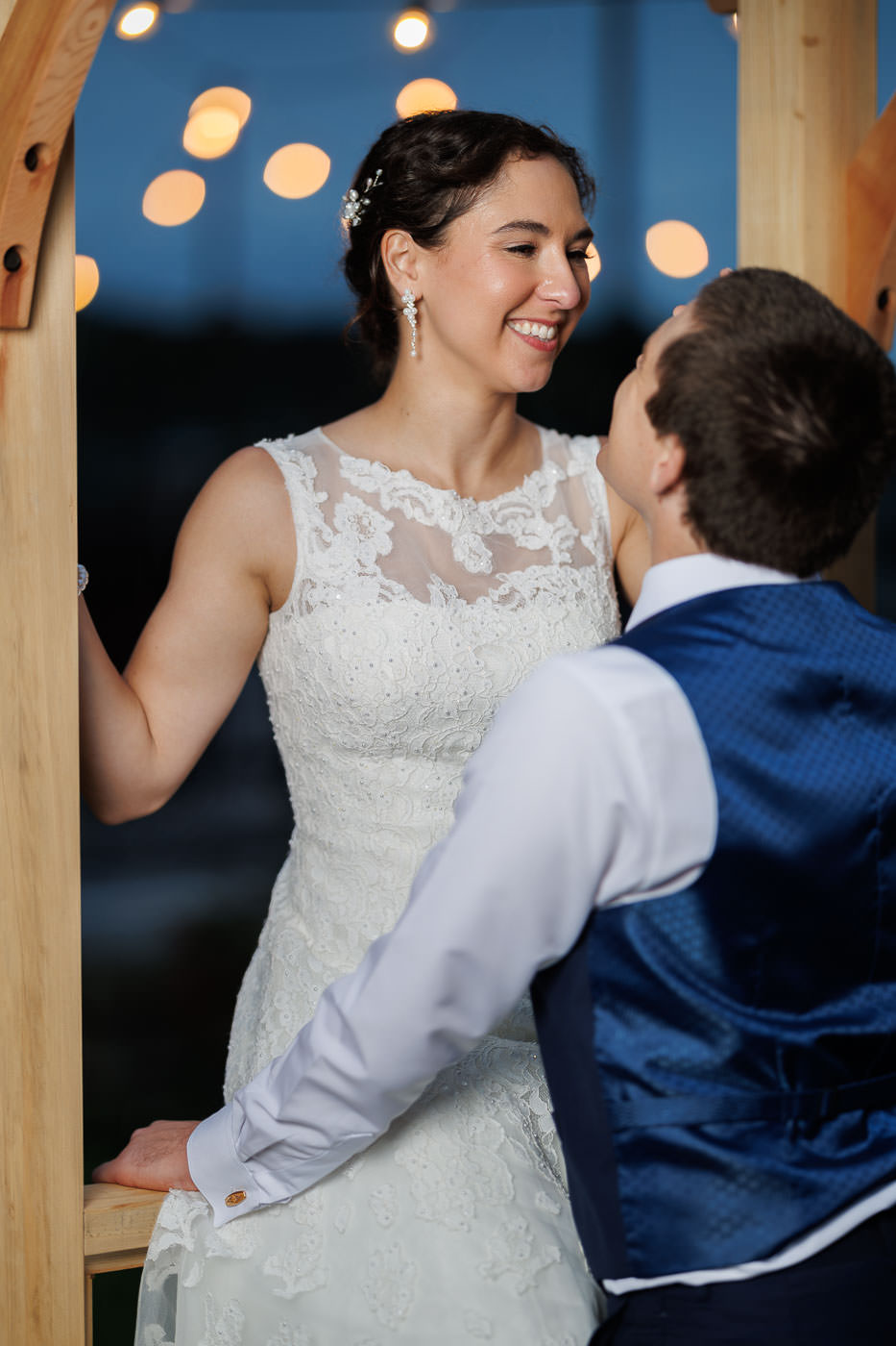 Lake house guest cottages wedding-20 A bride and groom share a moment under wooden archway lights at Bliss Farm, with the bride in a lace dress and the groom in a blue vest.