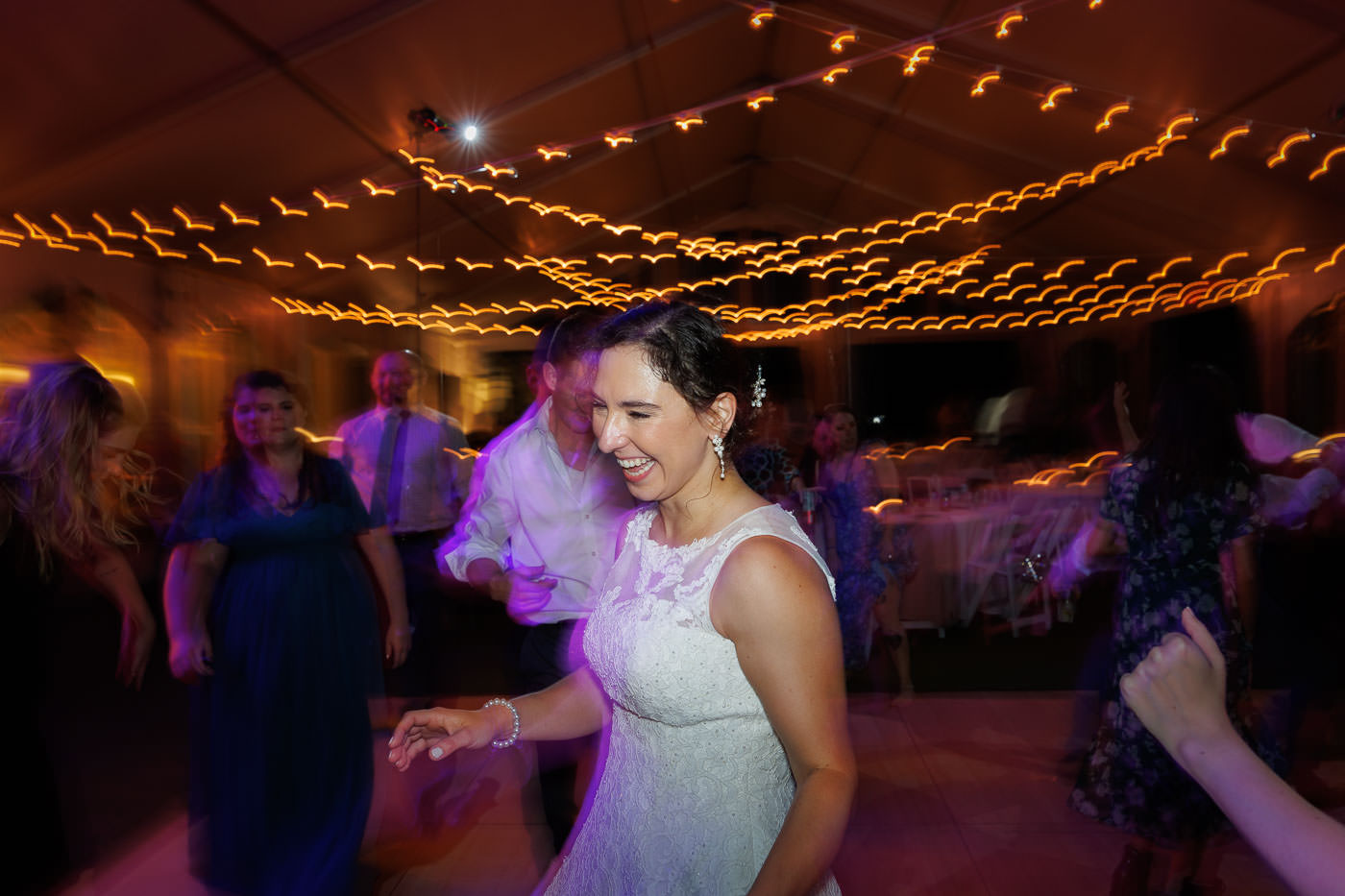 Lake house guest cottages wedding-23 At Bliss Farm, a bride in a white dress dances joyfully under string lights, surrounded by guests at the reception. Motion blur captures the lively atmosphere of this unforgettable wedding celebration.
