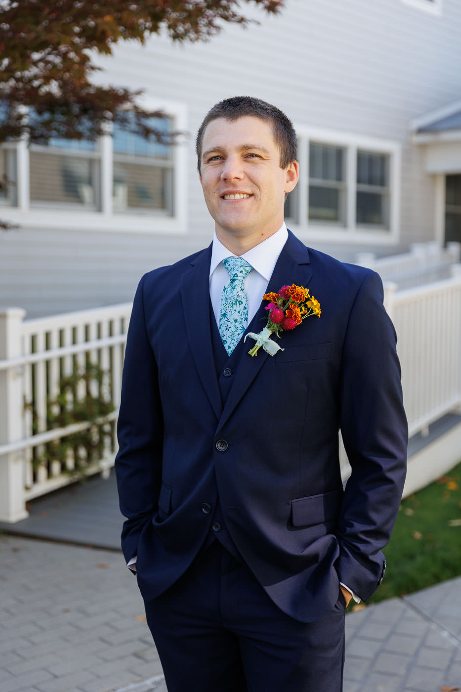 Lake house guest cottages wedding-4 A person in a dark suit with a floral boutonniere stands outdoors, exuding elegance at Bliss Farm, framed by a white fence and building.