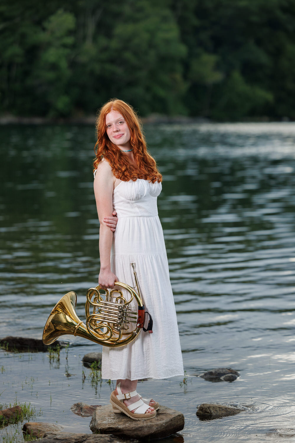 Person with long red hair in a white dress stands on a rock by a lake, holding a French horn. Trees are in the background.