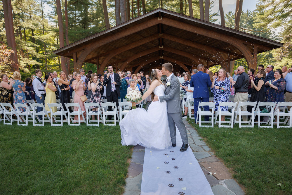 A bride and groom kiss at an outdoor wedding ceremony, surrounded by guests clapping and smiling under a wooden pavilion.