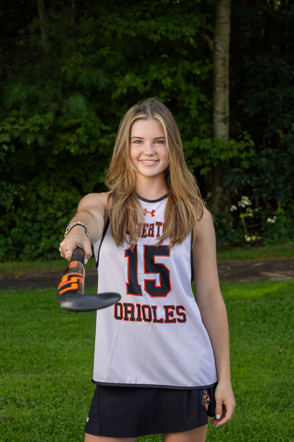 A young woman in a "Orioles" sports jersey holds a field hockey stick towards the camera, standing on grass with trees in the background.