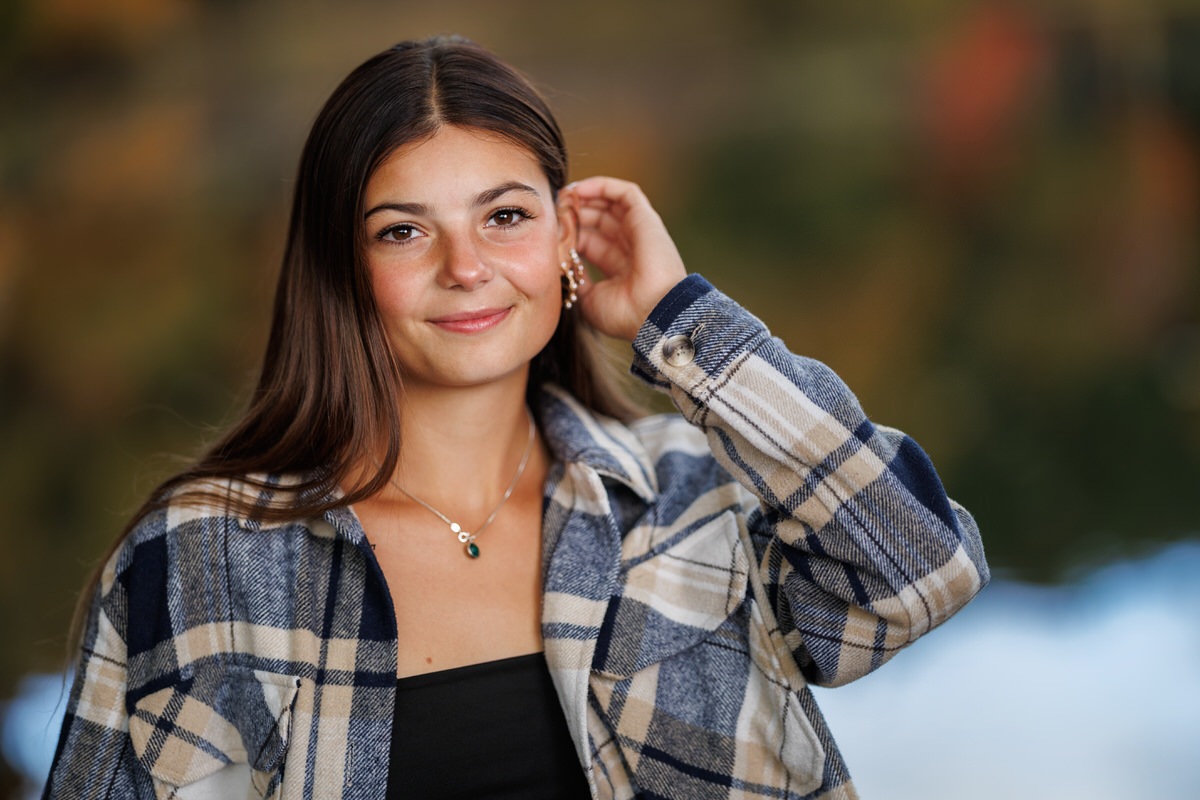 A high school senior girl with long hair wearing a plaid shirt and a black top adjusts her earring while smiling at the camera. The background is blurred.