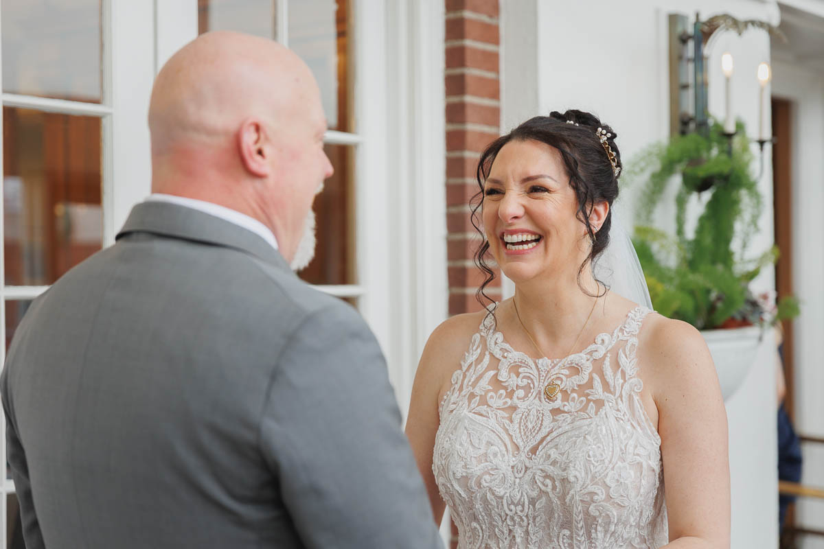 Hotel Northampton Wedding-10 A bride in a white lace dress laughs with a bald man in a gray suit, the elegant backdrop of The Northampton Hotel Wedding adding to the joyous atmosphere indoors.