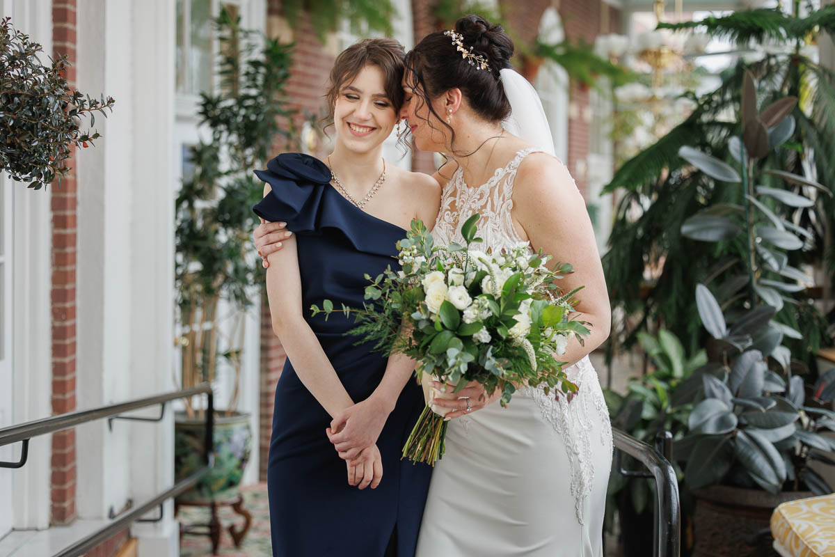 Hotel Northampton Wedding-11 Two women smiling at each other, one in a white wedding dress holding a bouquet, and the other in a navy blue dress. They stand in an elegant indoor setting at The Northampton Hotel Wedding, surrounded by charming brick walls and lush plants.