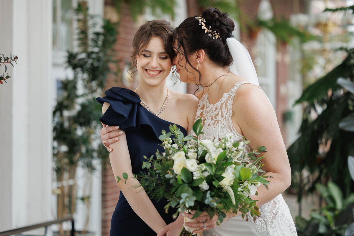 Hotel Northampton Wedding-12 At The Northampton Hotel wedding, a bride in a white lace gown clutches her bouquet while embracing a woman in a navy dress. Surrounded by lush greenery and the charm of an exposed brick wall, their joy illuminates the room.