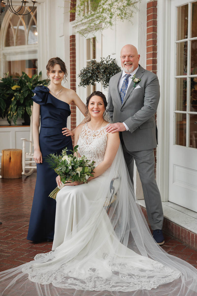 Hotel Northampton Wedding-13 A bride in a white dress sits holding a bouquet, flanked by a man in a gray suit and a woman in a navy dress, posing indoors at The Northampton Hotel Wedding. Plants and French doors elegantly frame this tender moment.