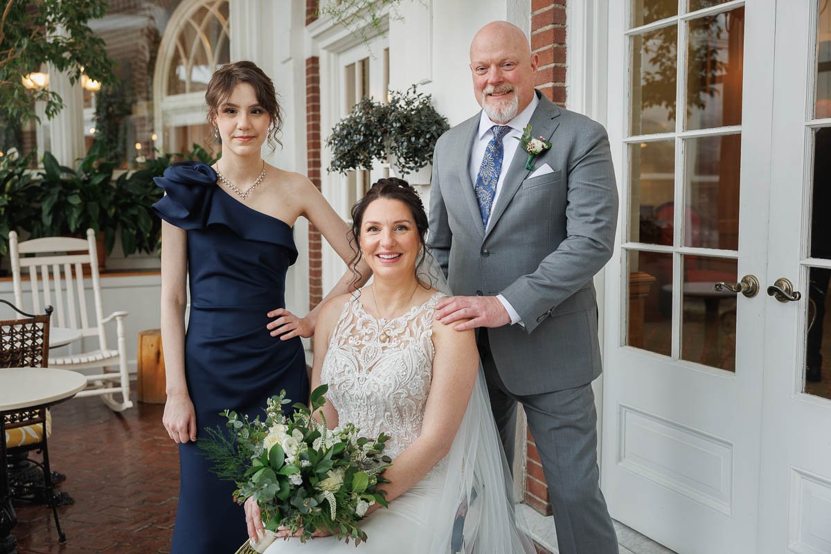Hotel Northampton Wedding-15 At The Northampton Hotel Wedding, a bride in a white gown holds a bouquet, seated between a man in a suit and a woman in a dark blue dress, all framed by the venue's elegant brick walls and glass doors.