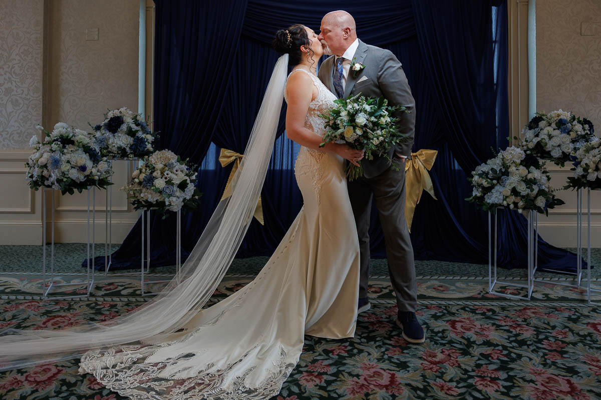 Hotel Northampton Wedding-16 A bride in a long dress and veil holds a bouquet while kissing a groom in a gray suit at The Northampton Hotel Wedding, surrounded by dark blue curtains and floral arrangements on both sides.