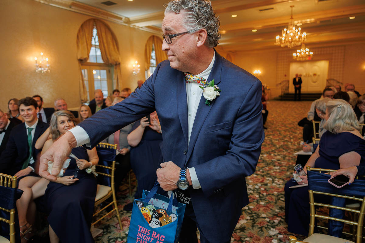 Hotel Northampton Wedding-19 At The Northampton Hotel Wedding, a man in a suit gracefully hands out items from a colorful bag to seated guests, all gathered in the elegant glow of the chandelier-lit room.