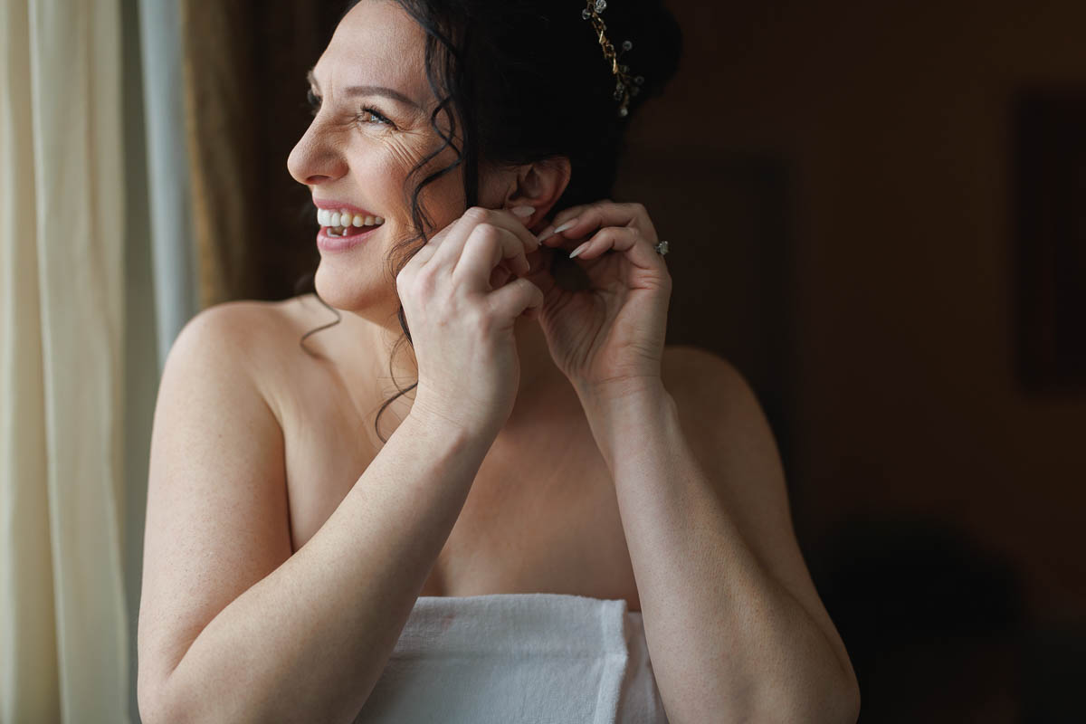 Hotel Northampton Wedding-2 Standing by a sunlit window, a woman smiles as she puts on earrings, preparing for her moment at The Northampton Hotel wedding.