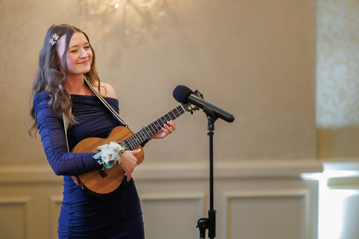 Hotel Northampton Wedding-20 At The Northampton Hotel Wedding, a woman in a blue dress plays a ukulele, her smile radiant as she stands by the microphone in the warmly lit room.