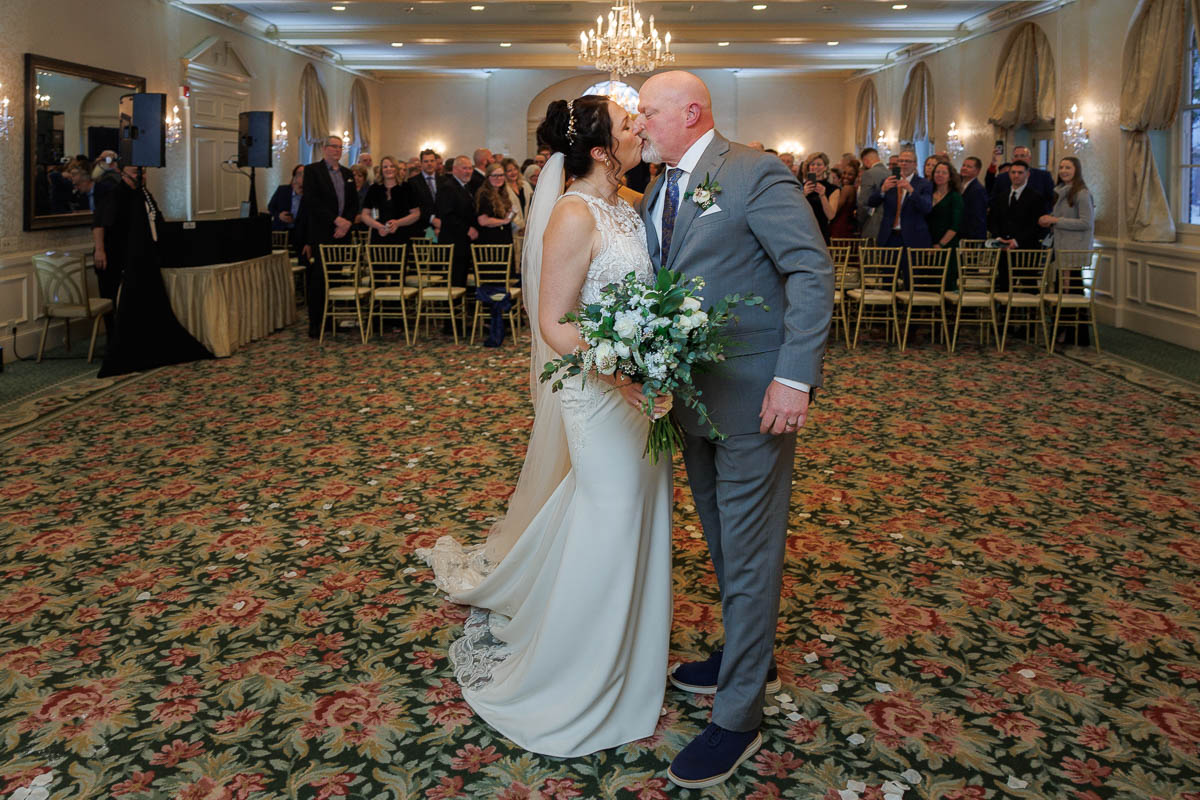 Hotel Northampton Wedding-22 A bride and groom share a kiss in a floral-carpeted room at their Northampton Hotel Wedding, with the seated audience beaming in the background.
