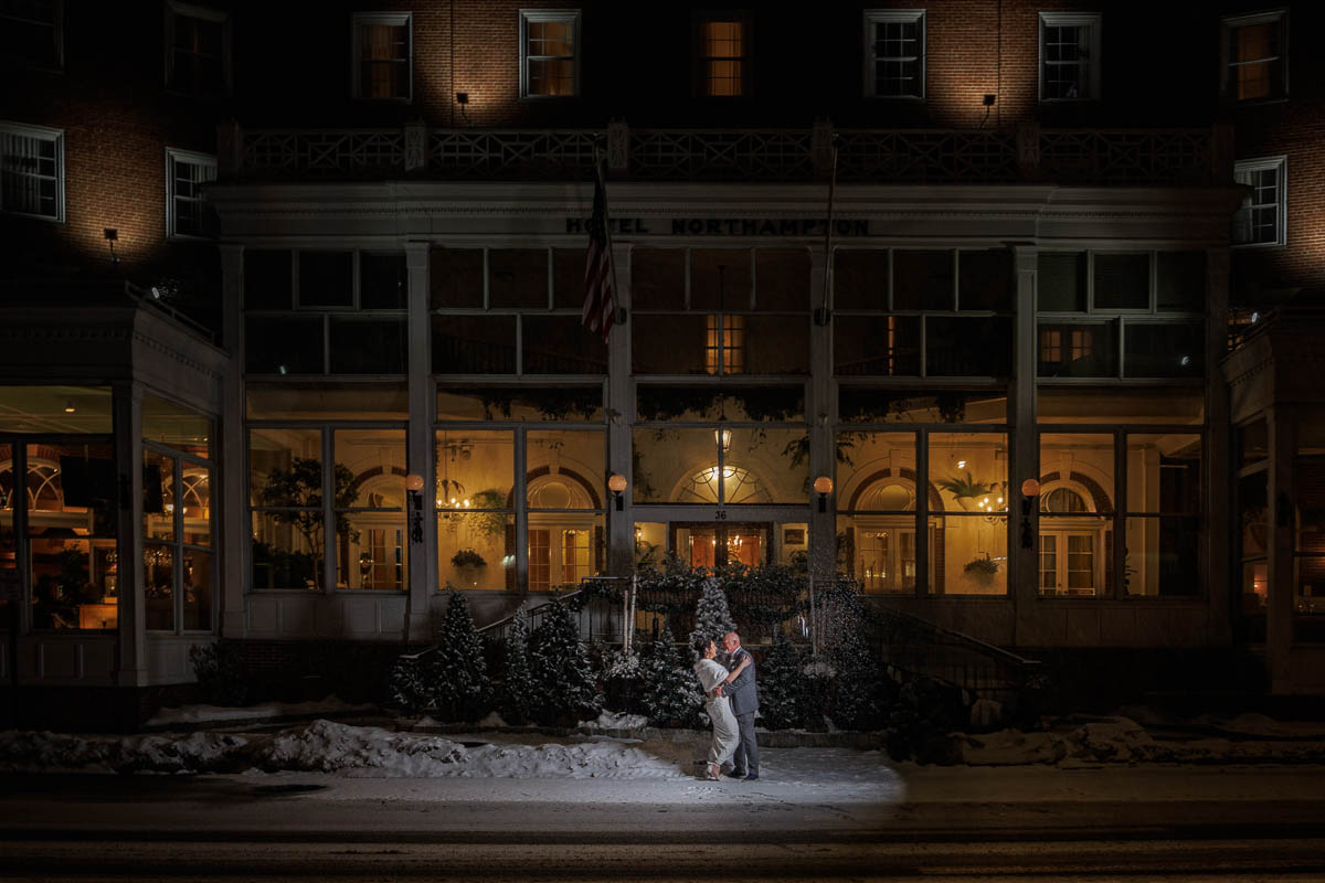 Hotel Northampton Wedding-24 A couple embraces on a snowy sidewalk at night in front of a lit building with the "Hotel Northampton" sign, capturing the magic of a Northampton Hotel wedding.