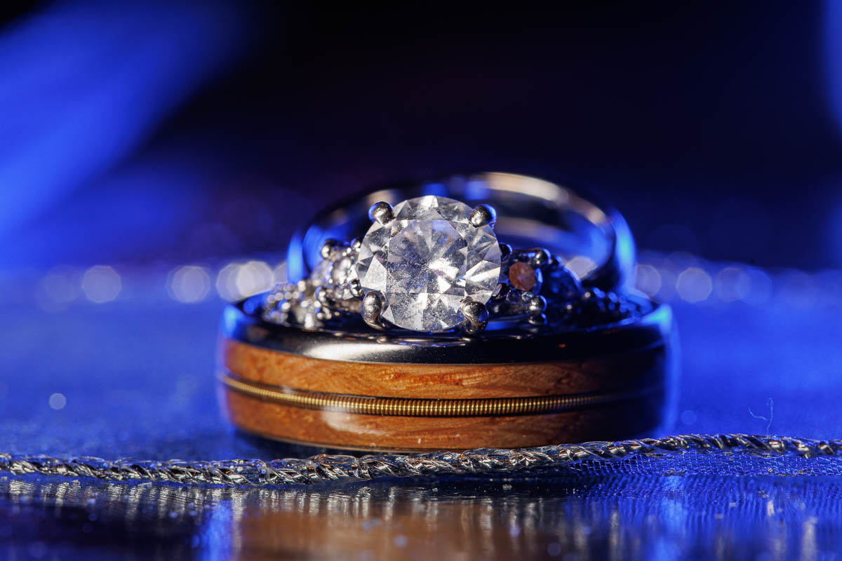 Hotel Northampton Wedding-25 A close-up of a diamond engagement ring and a wooden wedding band gleams on a reflective surface, set against a serene blue background. This stunning display evokes the elegance of The Northampton Hotel Wedding, where every detail sparkles with timeless romance.