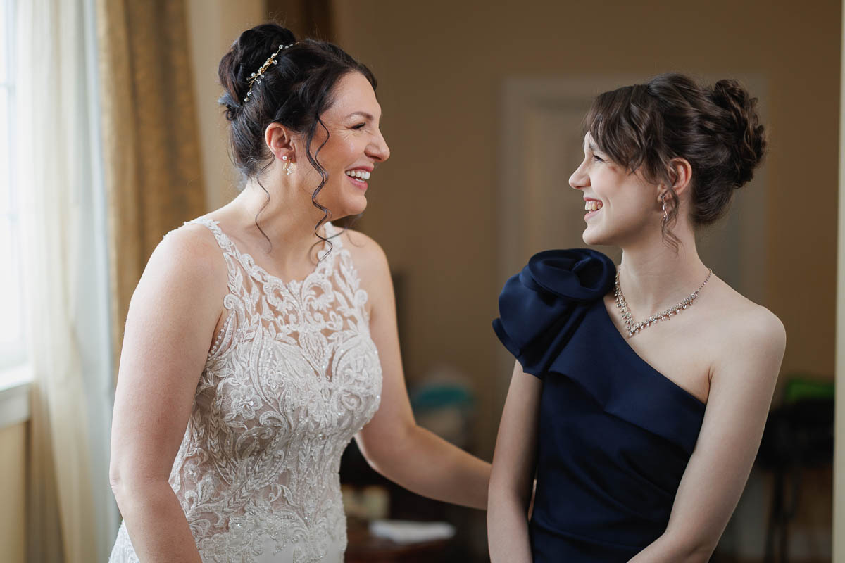 Hotel Northampton Wedding-3 Two women share a warm smile at The Northampton Hotel, one elegantly dressed in a white lace gown and the other in a chic navy blue one-shoulder dress, adding timeless charm to the indoor wedding ambiance.