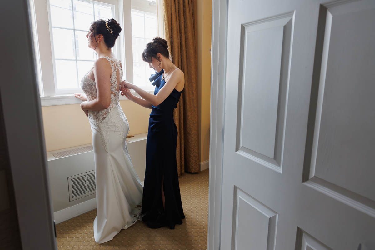Hotel Northampton Wedding-4 A woman in a white dress stands by the window at The Northampton Hotel, while another in a dark dress delicately adjusts the back of her gown.