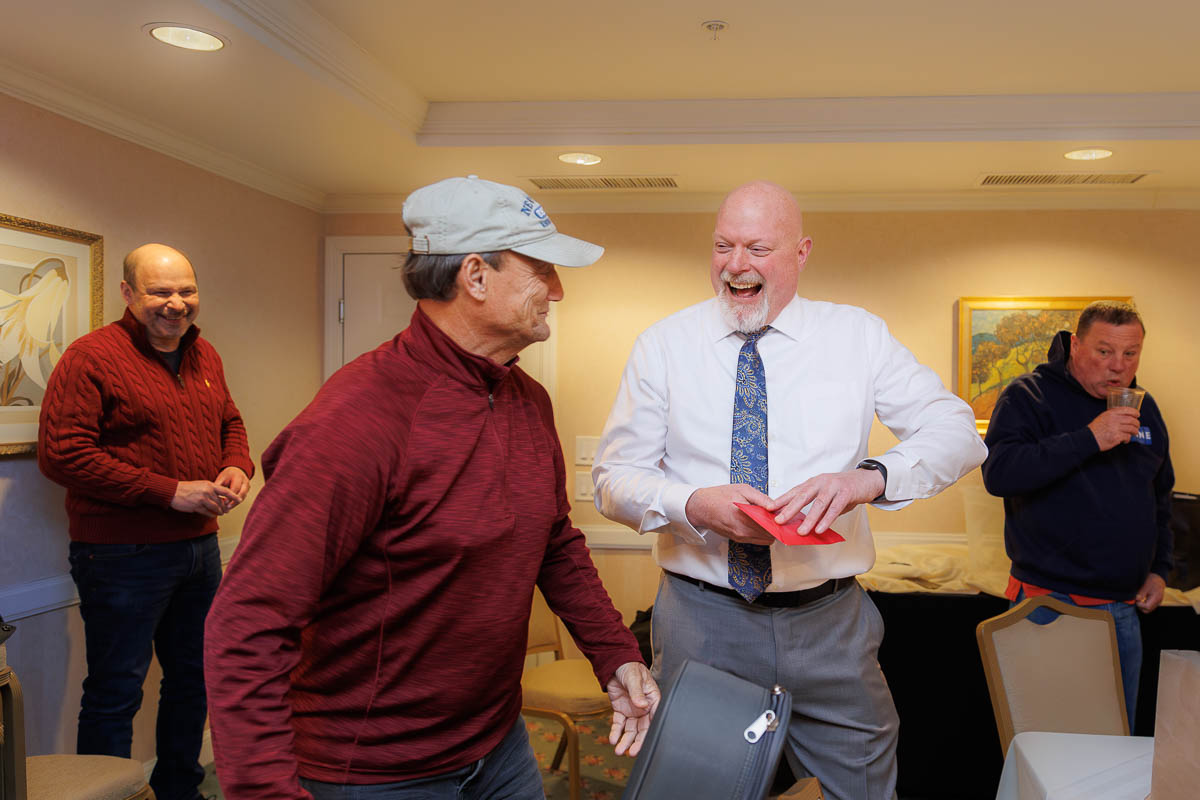 Hotel Northampton Wedding-5 In a cozy room at The Northampton Hotel Wedding, four men share a moment of laughter. One man, wearing a white shirt and tie, chuckles heartily while holding a red folder. The others stand nearby, smiling warmly in this joyous setting.
