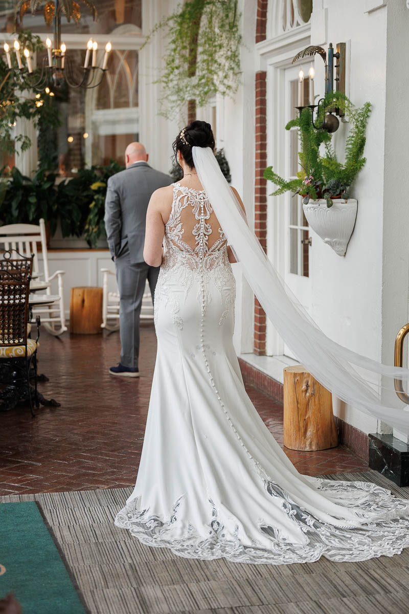 Hotel Northampton Wedding-8 A bride in a white gown with lace details and a long veil stands indoors at The Northampton Hotel, facing a man in a suit. Lush plants and natural light create an enchanting wedding setting.