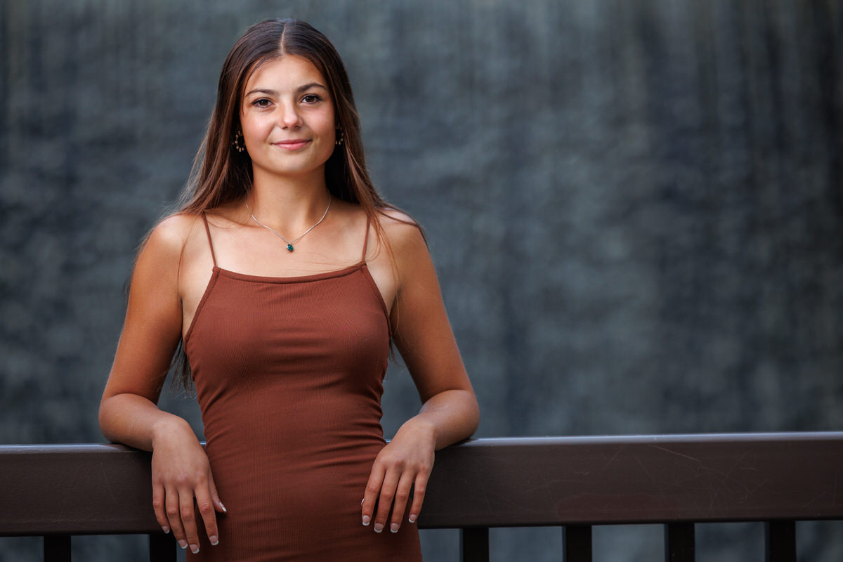 A woman in a brown dress stands and leans on a railing with a blurred waterfall backdrop.