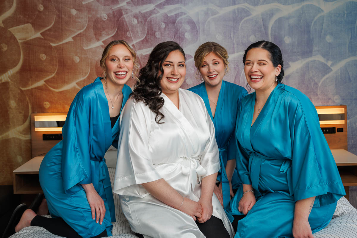 Maneeley’s banquet wedding-1-2 Four women in robes smile at the camera, capturing a moment reminiscent of Maneeley's Ct winter wedding photos. One wears a white robe, while the others don blue. They are seated on a bed with a patterned backdrop, radiating joy and warmth.