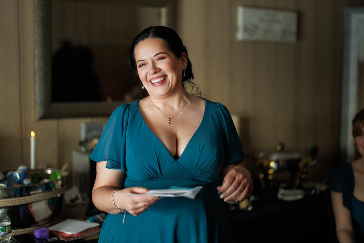 Maneeley’s banquet wedding-10 A woman in a teal dress stands indoors, holding papers and smiling. The room, reminiscent of Maneeley's Ct Winter Wedding Photos, has a mirror, candle, and various items on a table in the background.