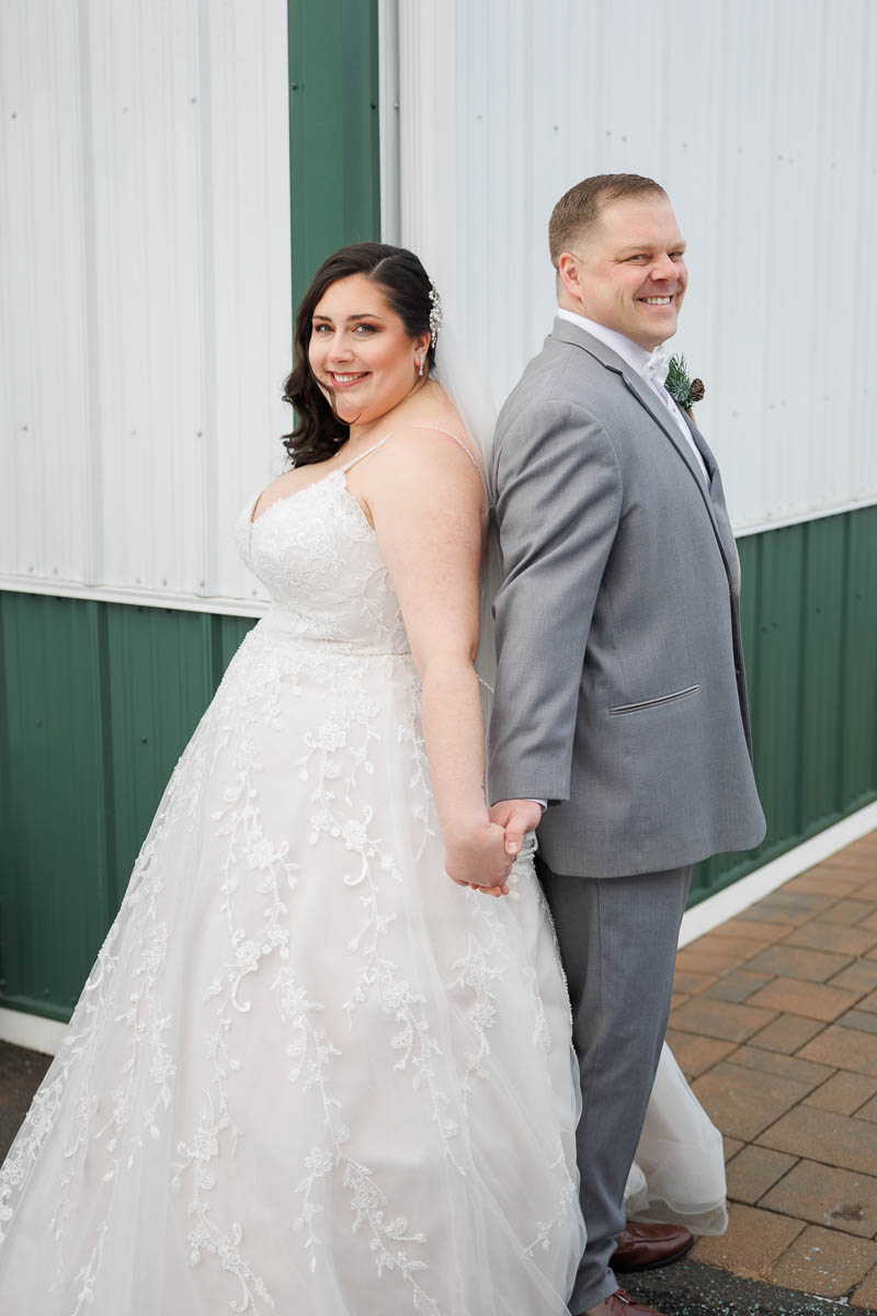 Maneeley’s banquet wedding-13 Bride and groom stand back to back holding hands, both smiling. Bride wears a lace gown; groom wears a gray suit. The backdrop of Maneeley's Ct captures the charm of a winter wedding, with the white and green building adding an elegant touch to their special photos.