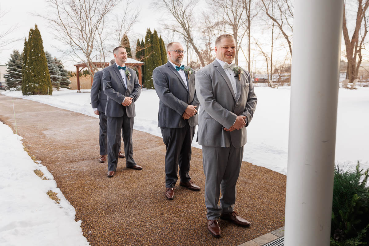 Maneeley’s banquet wedding-14 Four men in suits stand in a line outdoors on a snowy day, capturing the elegance of Maneeley's Ct winter wedding photos beside the pathway.