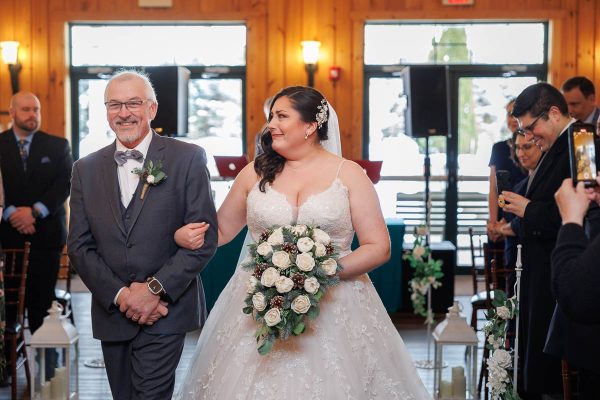 Bride walking down the aisle with an older man in a suit inside the wooden venue at Maneeley's Ct. She holds a bouquet of white flowers, while guests stand and watch, capturing perfect winter wedding photos.