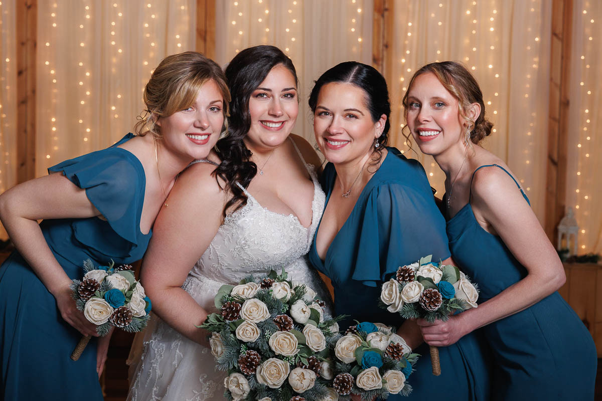 Maneeley’s banquet wedding-17 Bride in a white gown and three bridesmaids in blue dresses pose together holding bouquets, framed by the enchanting glow of string lights. This beautiful scene is perfect for capturing Maneeley's Ct Winter Wedding photos, creating memories that sparkle with warmth and joy.