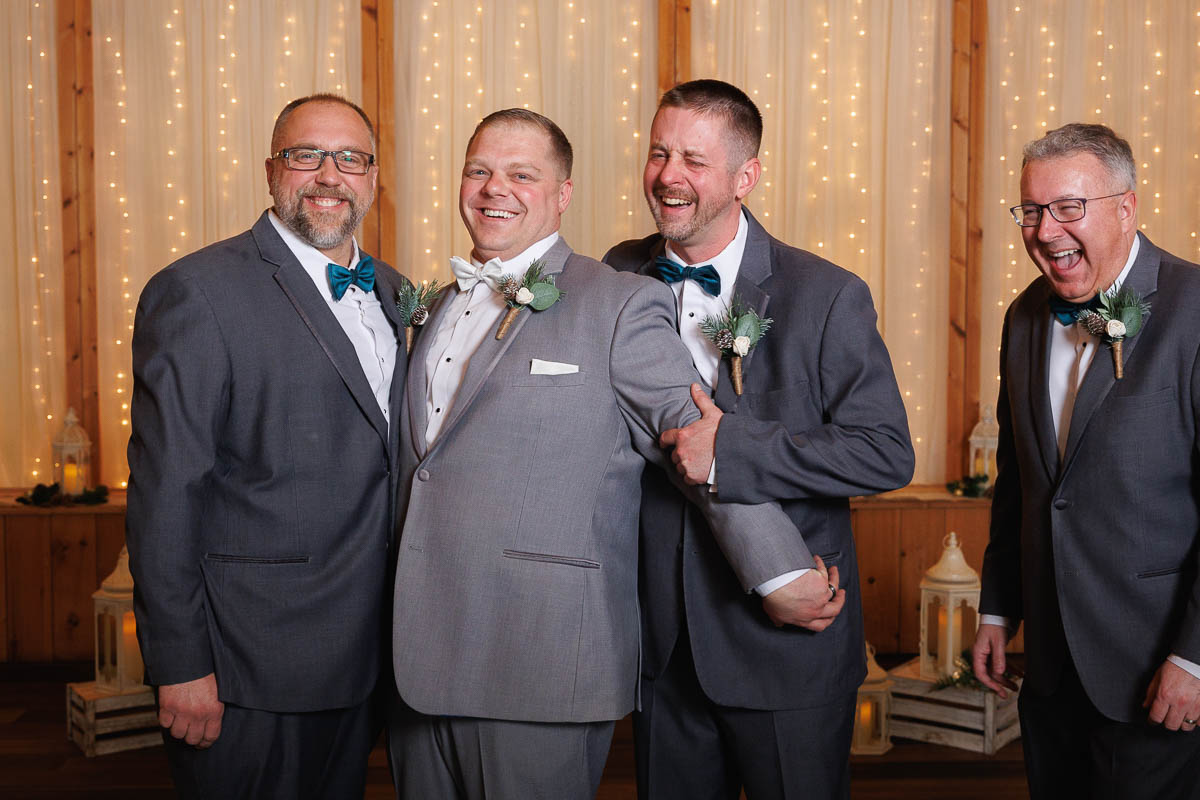 Maneeley’s banquet wedding-18 Four men in suits smile and pose together at Maneeley's Ct, with string lights twinkling, capturing the warmth of a winter wedding.