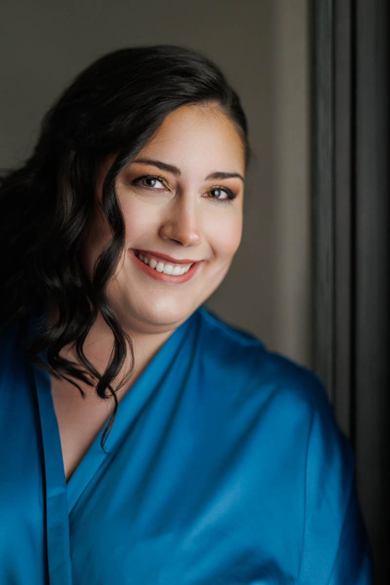 Maneeley’s banquet wedding-2-2 A woman with dark wavy hair smiles while wearing a bright blue robe, standing near a window, capturing the serene elegance seen in Maneeley's Ct Winter Wedding Photos.