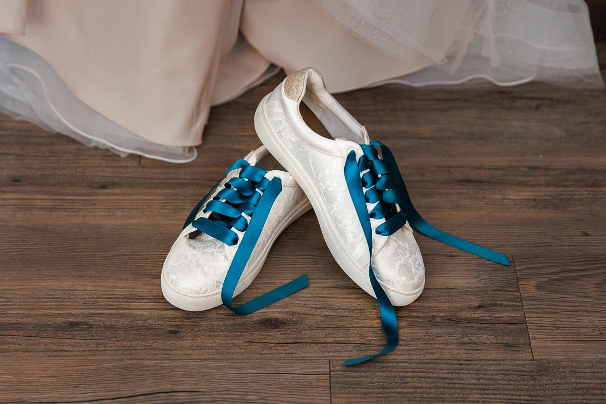 Maneeley’s banquet wedding-2 White sneakers with a lacy texture and teal satin laces rest elegantly on a wooden floor next to a light-colored garment, reminiscent of those Maneeley’s Ct winter wedding photos.