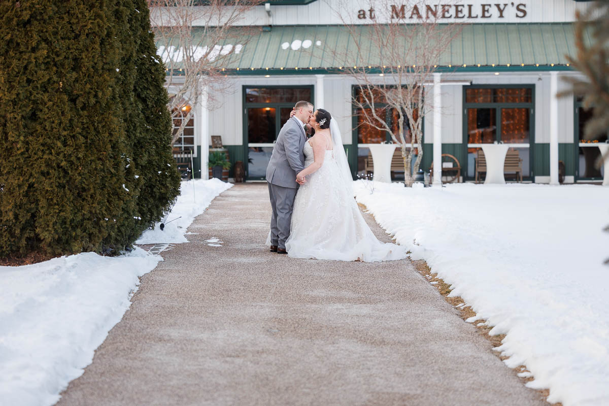 Maneeley’s banquet wedding-20 The bride and groom share a kiss on the snowy pathway at Maneeley's Ct, framed by a building with green trim and surrounded by trees, capturing enchanting winter wedding photos.