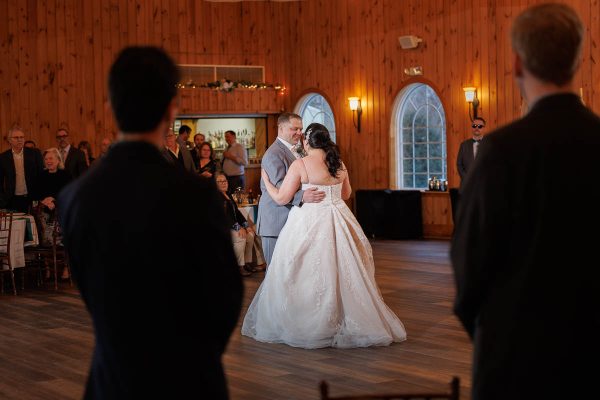 In the cozy, wooden-paneled room at Maneeley's Ct, a couple gracefully spins under the warm glow of lights. It's a winter wedding scene to remember, with guests enchanted by their dance. In the foreground, two figures silently capture the magic with their cameras. .