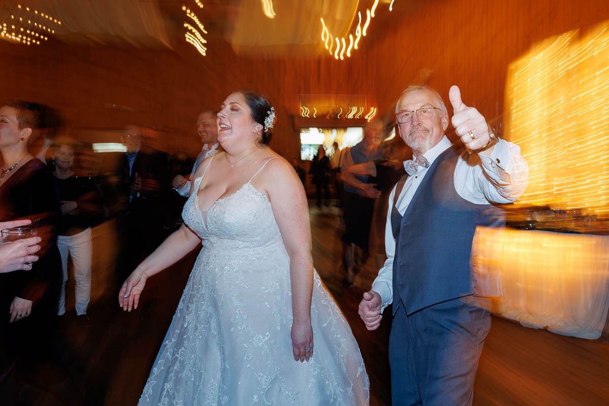 Maneeley’s banquet wedding-24 A bride in a white gown and a man in a suit are on the dance floor at Maneeley's Ct. The man is giving a thumbs up, capturing the joy of their winter wedding. The background is blurred with twinkling lights visible, adding to the enchanting atmosphere.