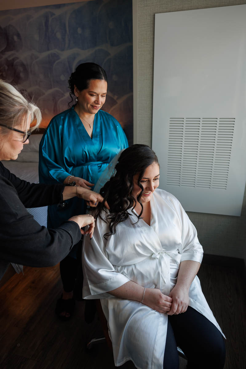 Maneeley’s banquet wedding-3 A woman in a white robe sits smiling while a stylist meticulously curls her hair, preparing for the Maneeley's Ct winter wedding photos, as another woman in a blue robe stands nearby, ready to assist.