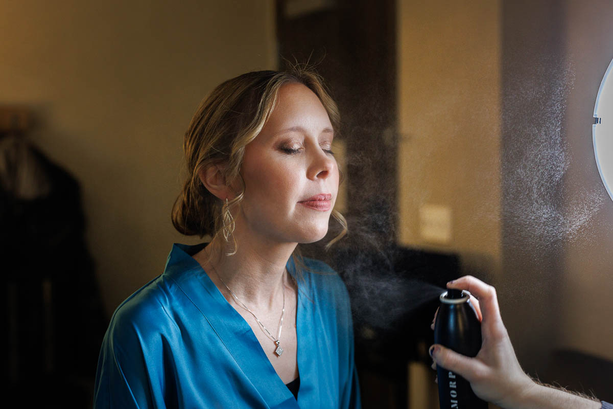 Maneeley’s banquet wedding-4 A woman in a blue robe has her eyes closed while getting misted with a spray from a can, evoking the serene elegance of Maneeley's Ct winter wedding photos, with clothes hanging gently in the background.