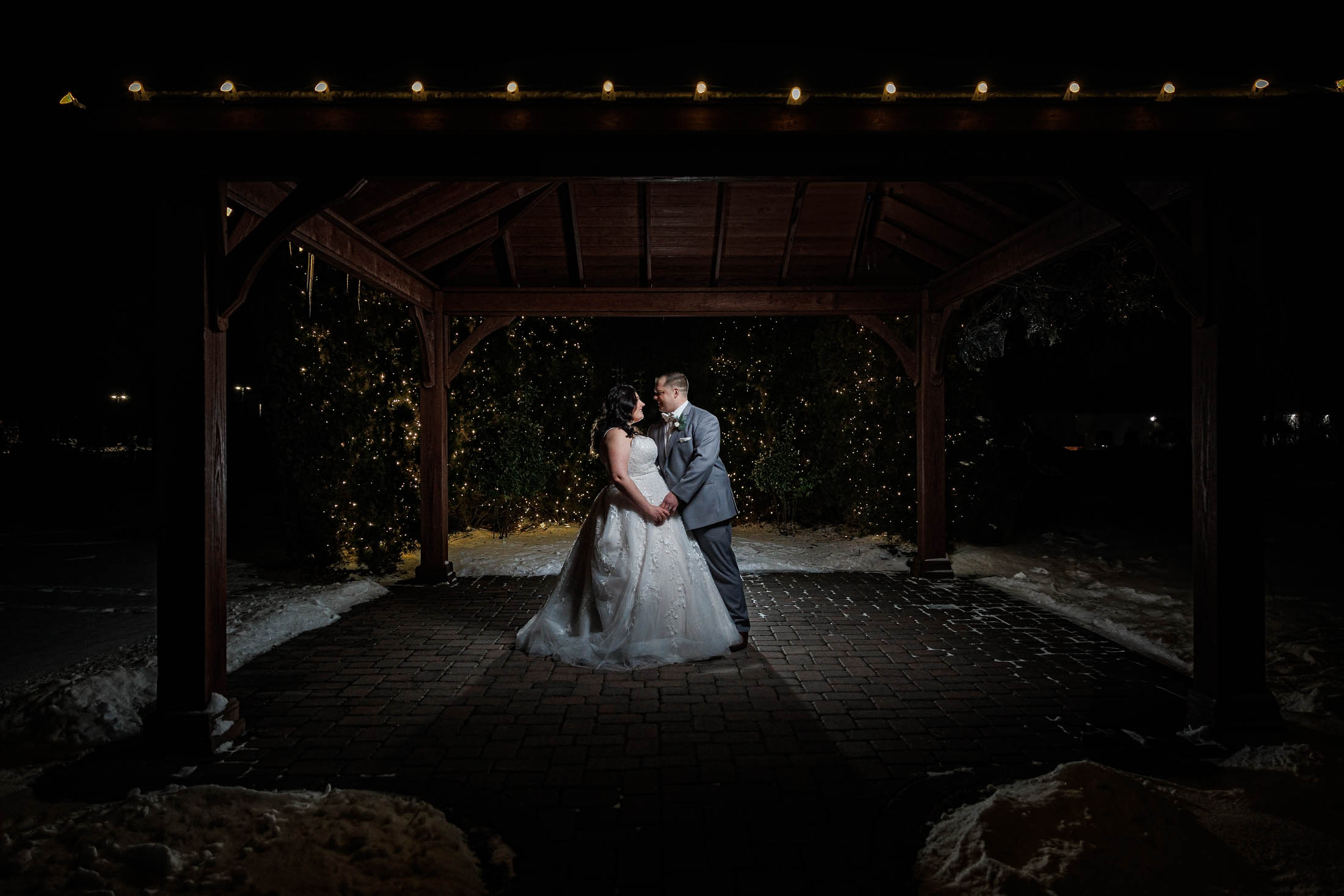 At Maneeley's Ct, the bride and groom stand under a lit wooden pavilion, their winter wedding photos capturing the enchanting scene surrounded by snow.