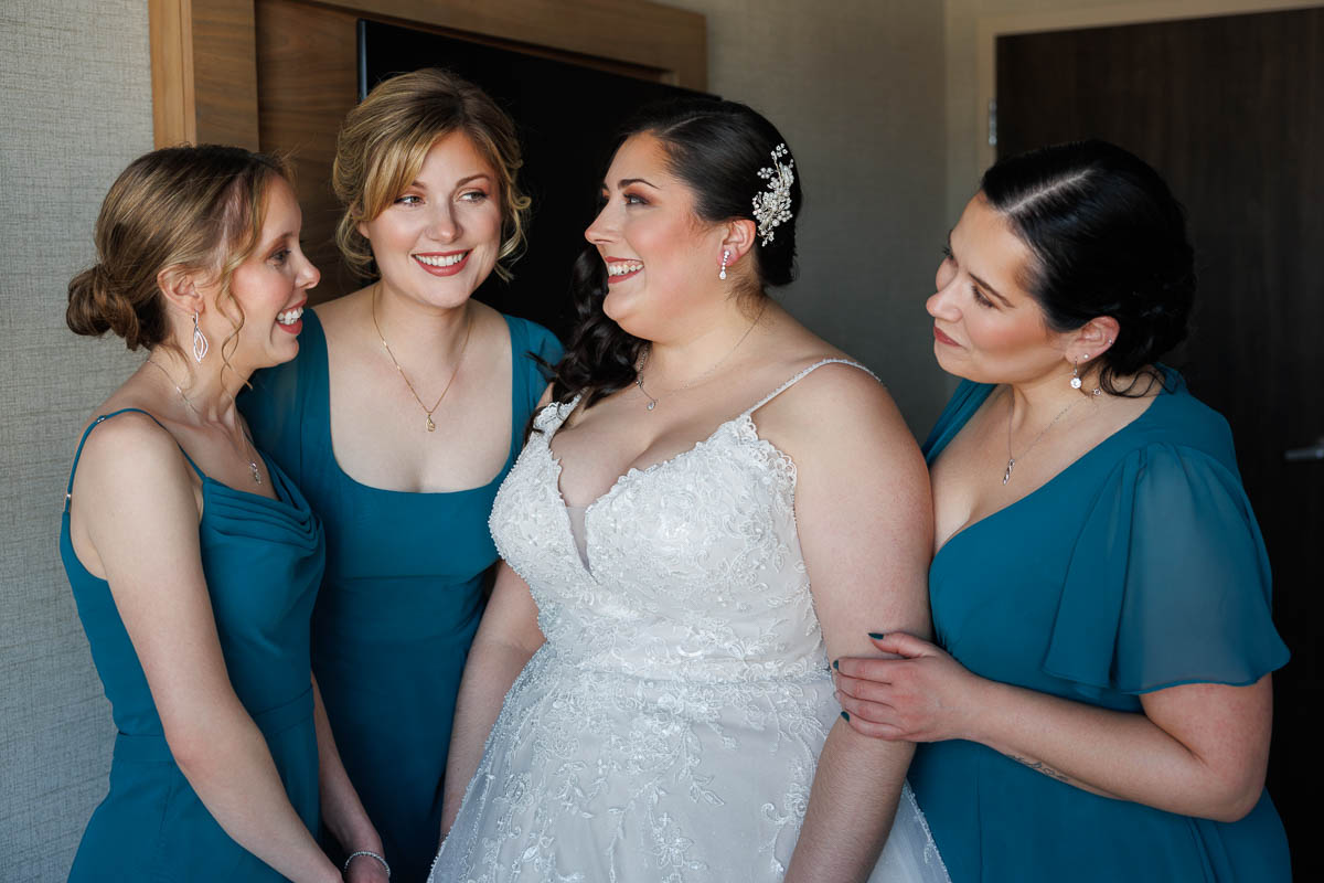 Maneeley’s banquet wedding-8 In this heartwarming Maneeley's Ct Winter Wedding photo, the bride beams in her white lace gown and hairpiece, holding hands with one of her three bridesmaids dressed in elegant teal. Their joy is palpable as they stand indoors, capturing a moment of pure happiness.