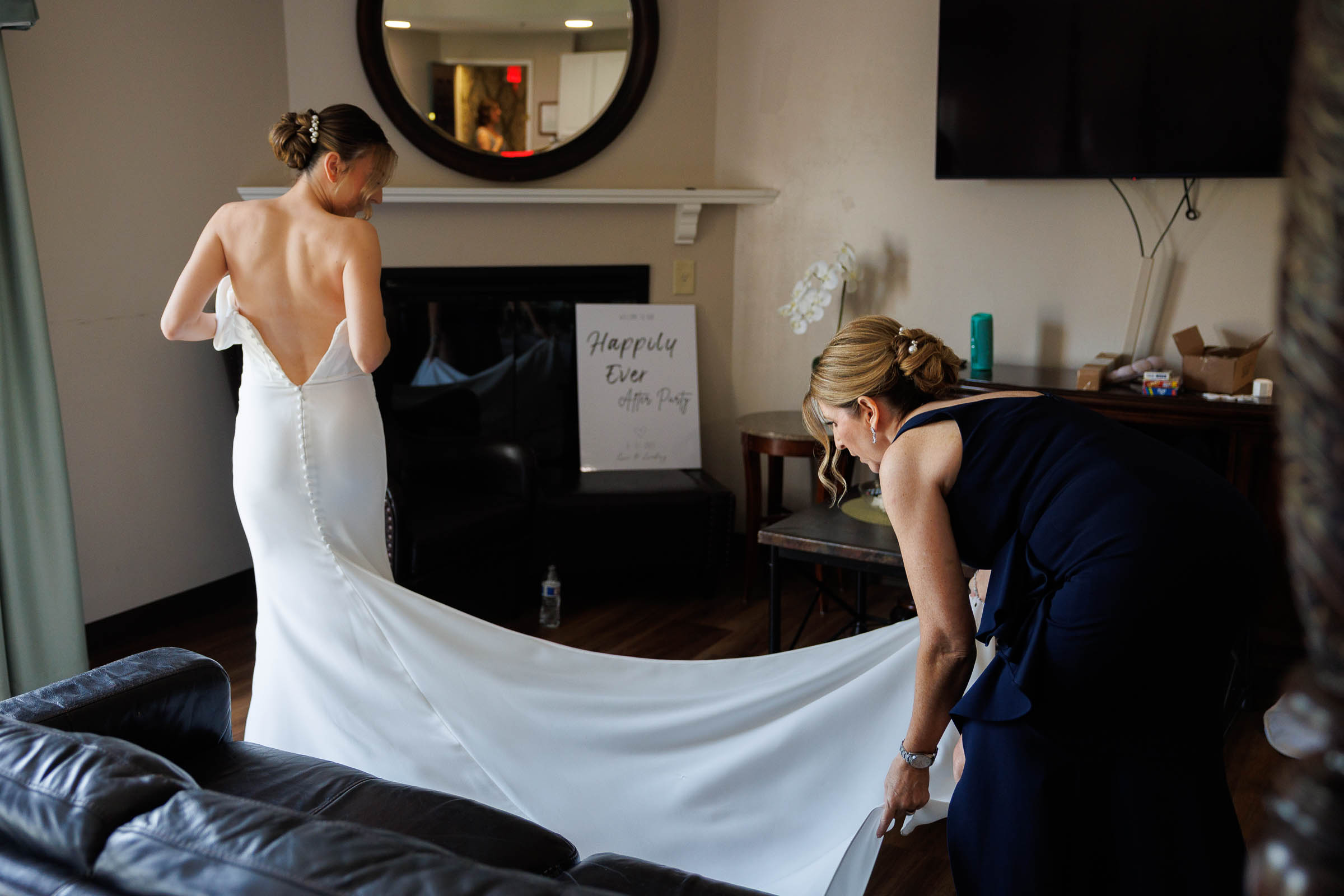 Log Cabin Wedding Photos Holyoke-10 A bride in a white dress stands as another woman adjusts her train in a living room with a "Happily Ever After Party" sign—capturing one of many unforgettable Log Cabin Holyoke wedding photos.