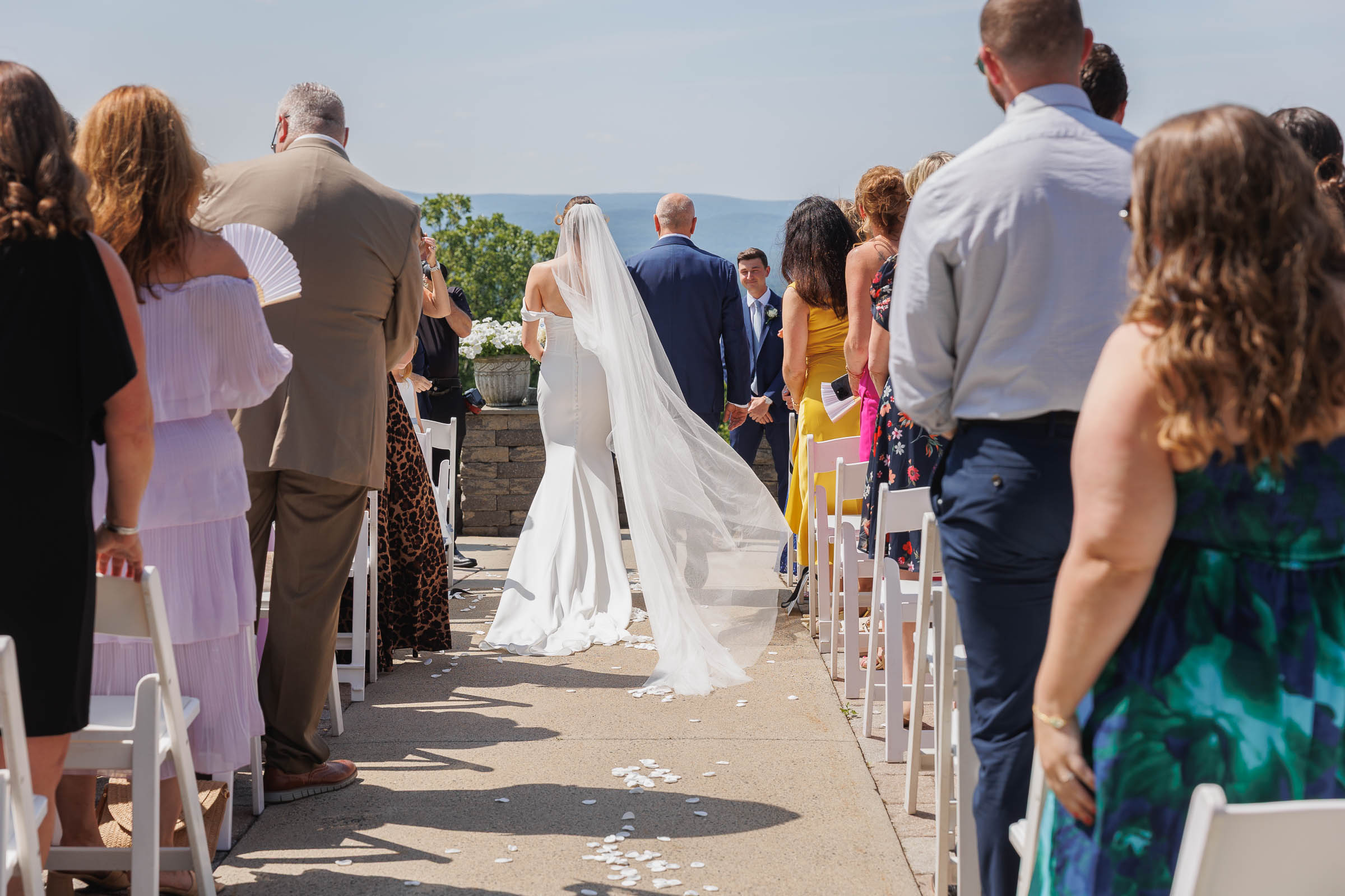 Log Cabin Wedding Photos Holyoke-15 A bride in a white dress and veil walks down an outdoor aisle lined with seated guests, towards a groom at the altar on a sunny day—perfect for The Log Cabin Holyoke wedding photos.