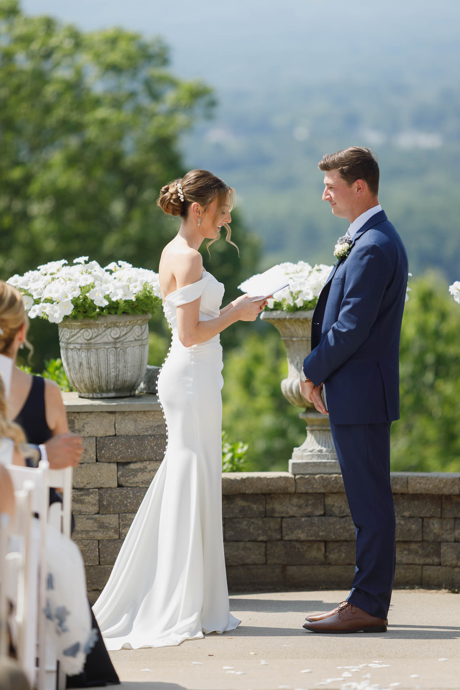 Log Cabin Wedding Photos Holyoke-17 A bride in a white dress and a groom in a navy suit stand facing each other outdoors at The Log Cabin Holyoke, with lush greenery and white flowers creating the perfect backdrop for unforgettable wedding photos.
