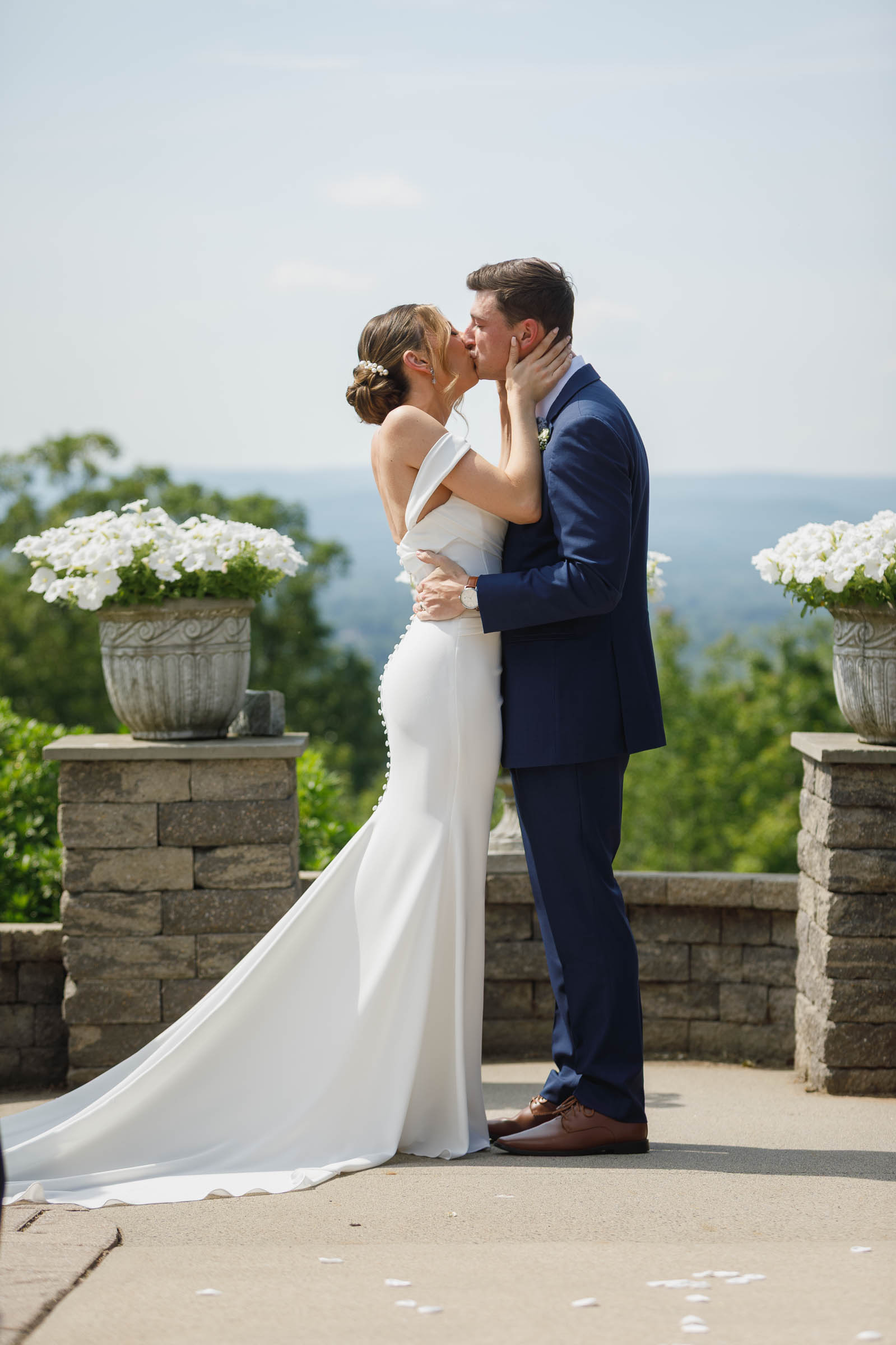 Log Cabin Wedding Photos Holyoke-19 A bride and groom kiss outdoors in front of stone planters with white flowers, greenery, and hills in the background—capturing the romance of The Log Cabin Holyoke wedding photos.
