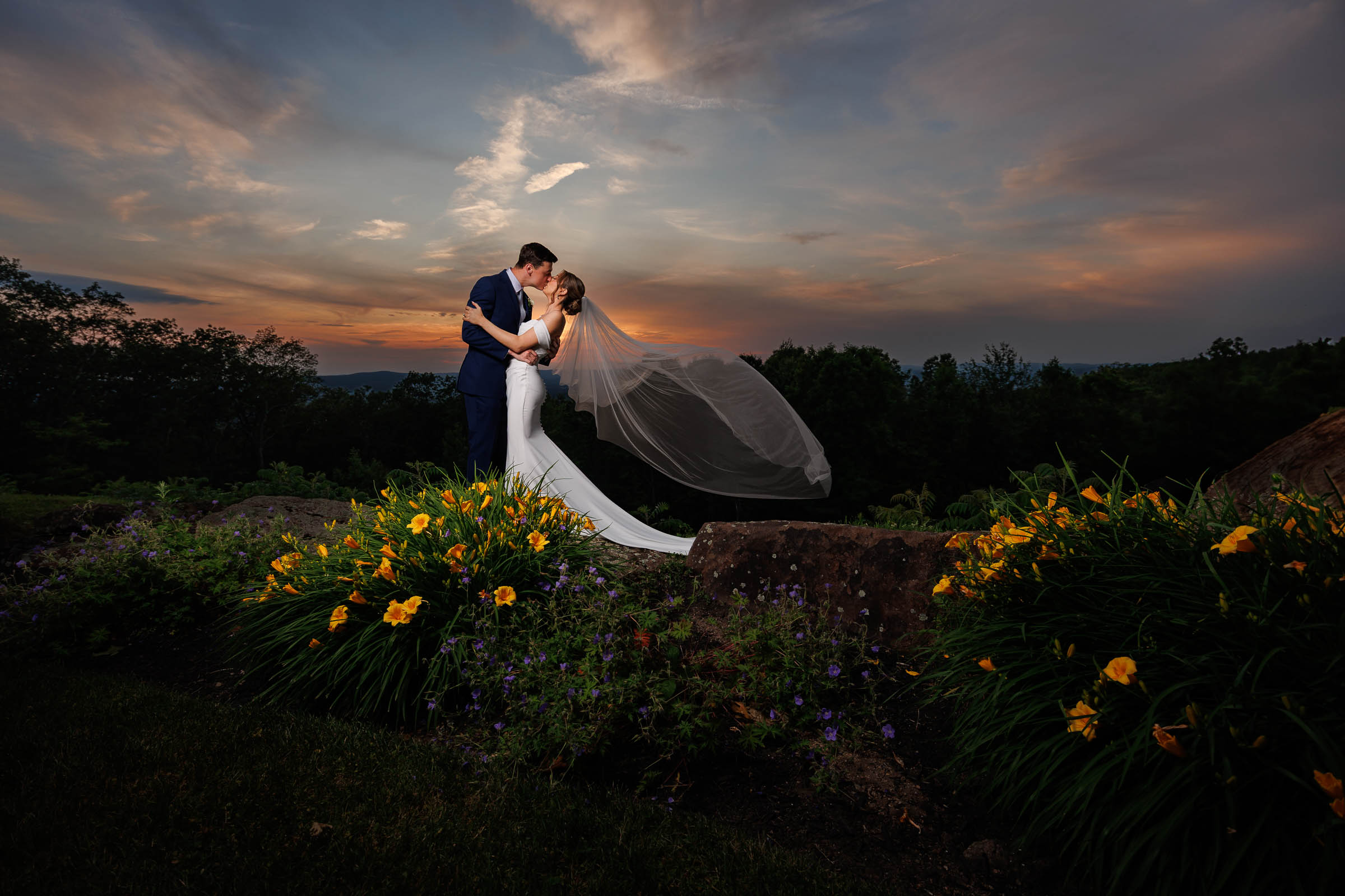 A bride and groom embrace outdoors at sunset, surrounded by yellow flowers, with the bride's veil flowing behind her in this beautiful The Log Cabin Holyoke wedding photo.
